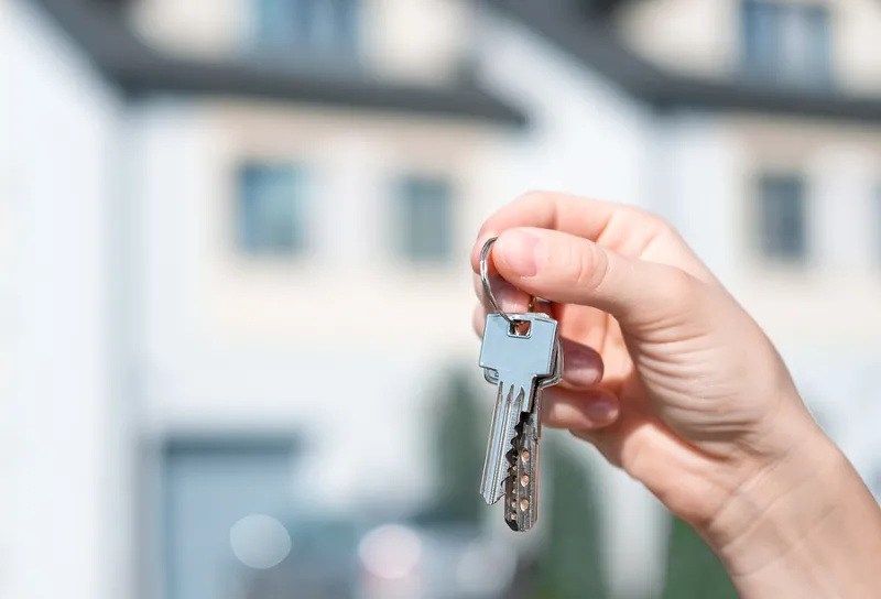 Holding House Keys Front Of A New Home. Keys On The Background O